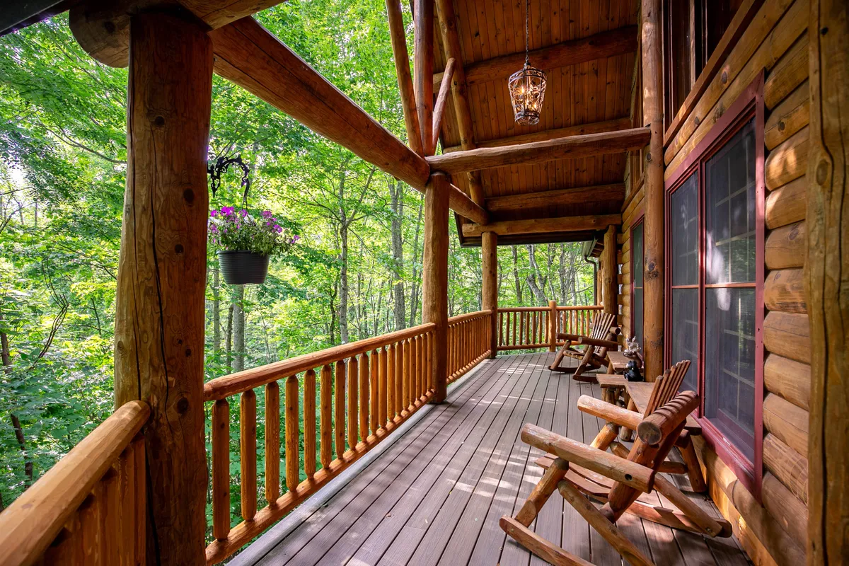 An exterior porch with wooden rocking chairs, a hanging flower pot, natural wood railings, and the log structure of the home visible. The scene conveys a peaceful outdoor atmosphere, ideal for relaxing, reading, or enjoying the sounds and views of nature.