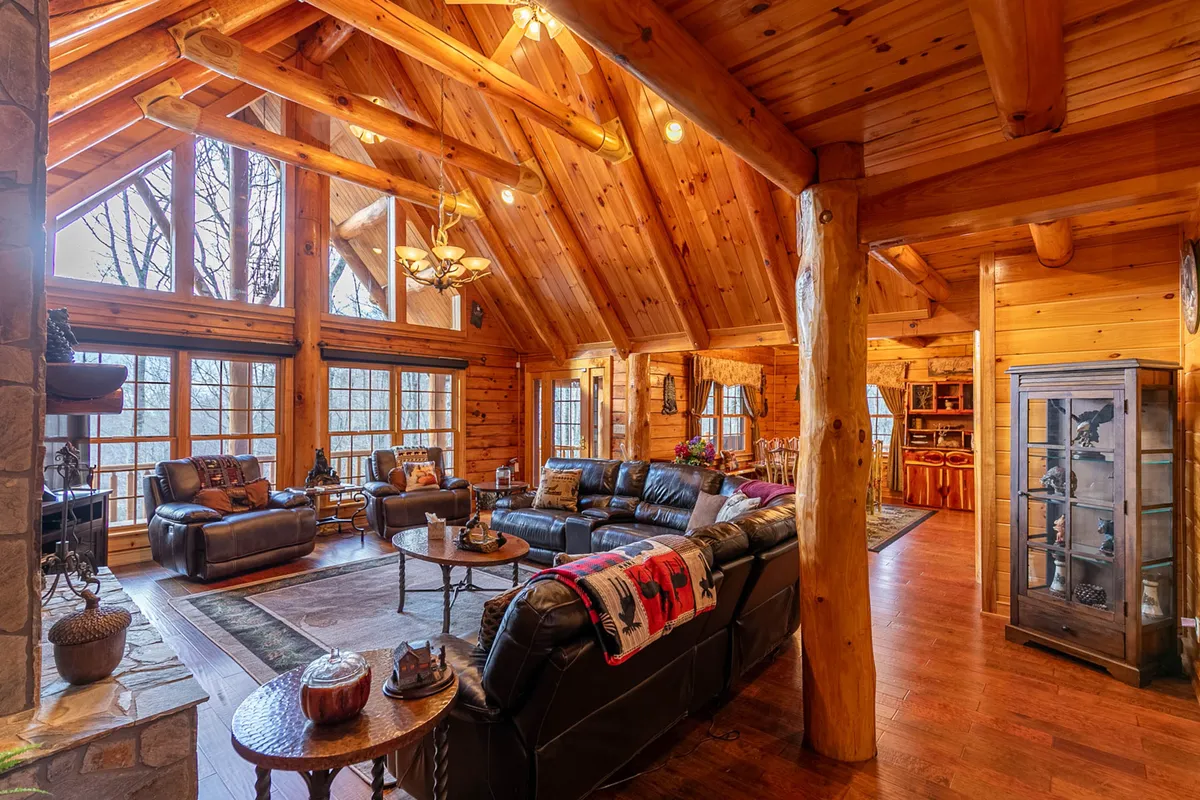The image shows a cozy living area with large windows overlooking the mountains, wooden beams overhead, and plush leather sofas arranged around a central coffee table. The space exudes warmth and offers an intimate setting for conversation or relaxation while enjoying the surrounding natural beauty.