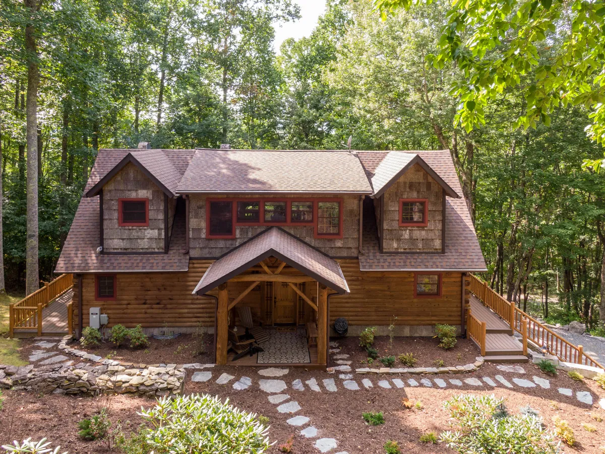 This exterior view highlights the log home's architectural character, with a cozy porch featuring seating, red-framed windows, and a stone pathway. The inviting outdoor space connects the home to the surrounding landscape, emphasizing comfort and family-friendly design.