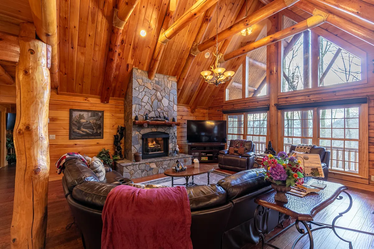This image captures a spacious great room featuring a stunning stone fireplace, exposed wooden beams, plush leather sofas, and large windows that frame scenic mountain views. Decorative accents, such as fresh flowers, add a touch of elegance to the rustic setting. The space feels open, warm, and perfect for gatherings.