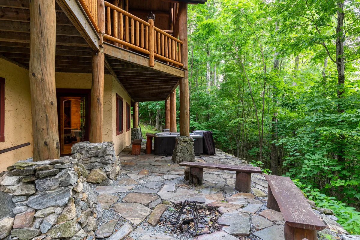 A cozy outdoor area featuring a stone fire pit with a wooden bench and a hot tub nearby, surrounded by nature and supported by sturdy wooden pillars. The space invites gatherings for marshmallow roasting, relaxation, and outdoor celebrations in a serene environment.