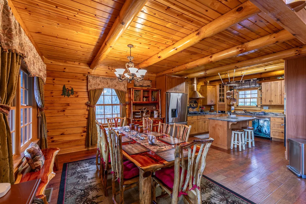A long wooden dining table sits under a rustic chandelier, adjacent to a chef’s kitchen with wooden cabinetry. The large windows allow natural light to flood the open space, creating a welcoming environment for dining, socializing, and celebrating with family and friends.