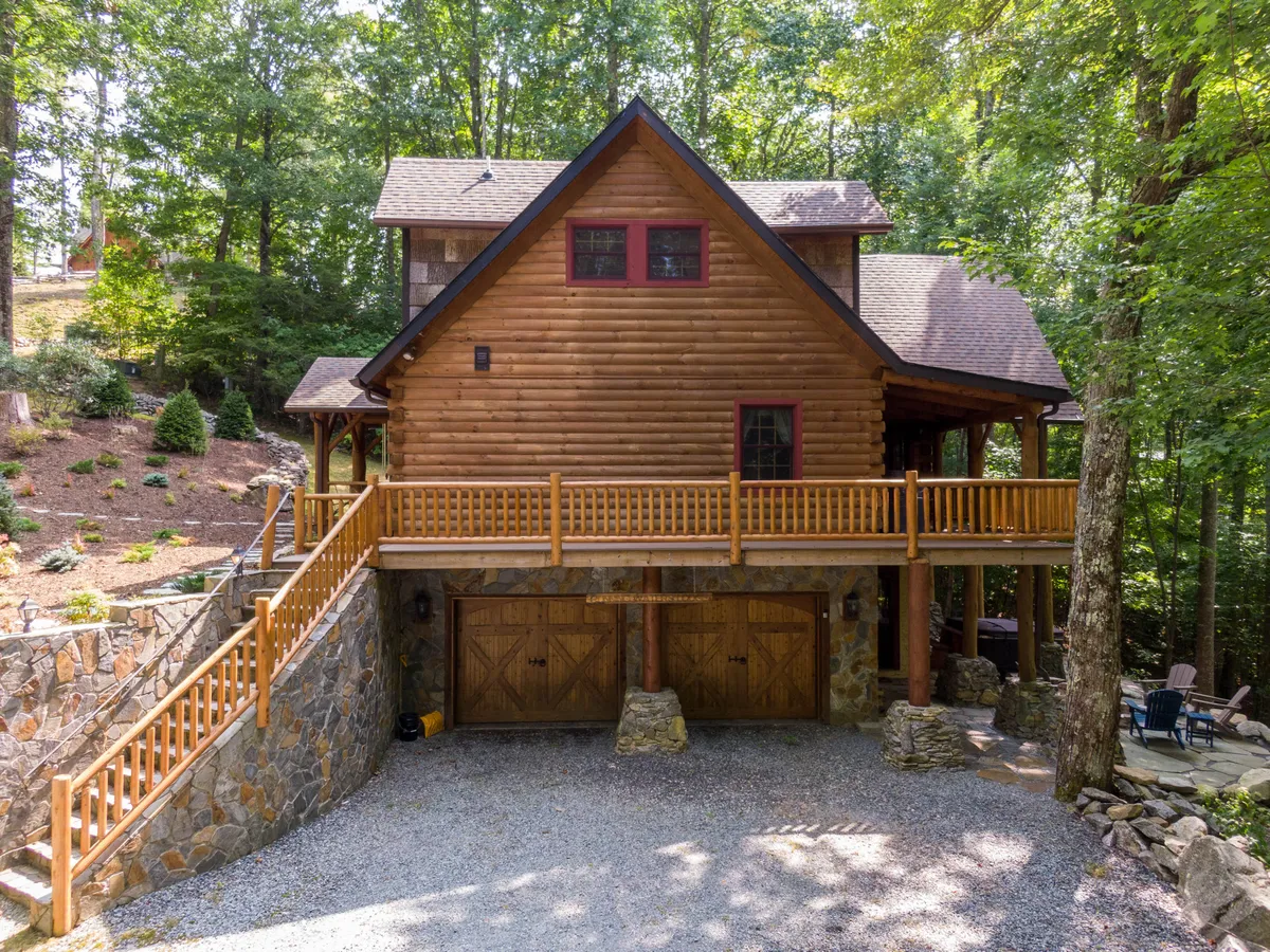 A cozy log cabin with a large deck is nestled among tall trees, featuring a stone foundation and surrounded by natural greenery. The inviting atmosphere suggests a direct connection to nature and easy access to outdoor activities like hiking and gathering.