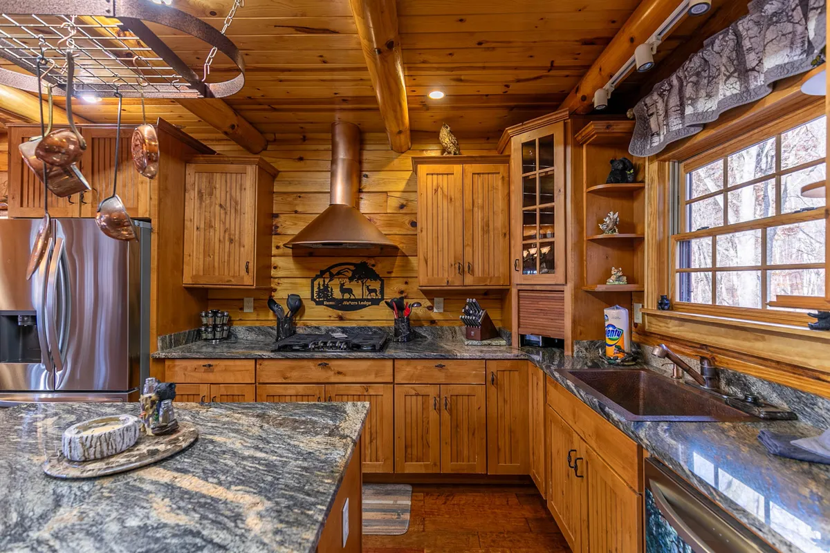 The kitchen features a beautiful granite countertop, warm wood cabinets, a prominent copper range hood, and a large window that fills the space with natural light. Decorative shelves add a homey, rustic touch, while the view to the outdoors evokes the mountain setting.