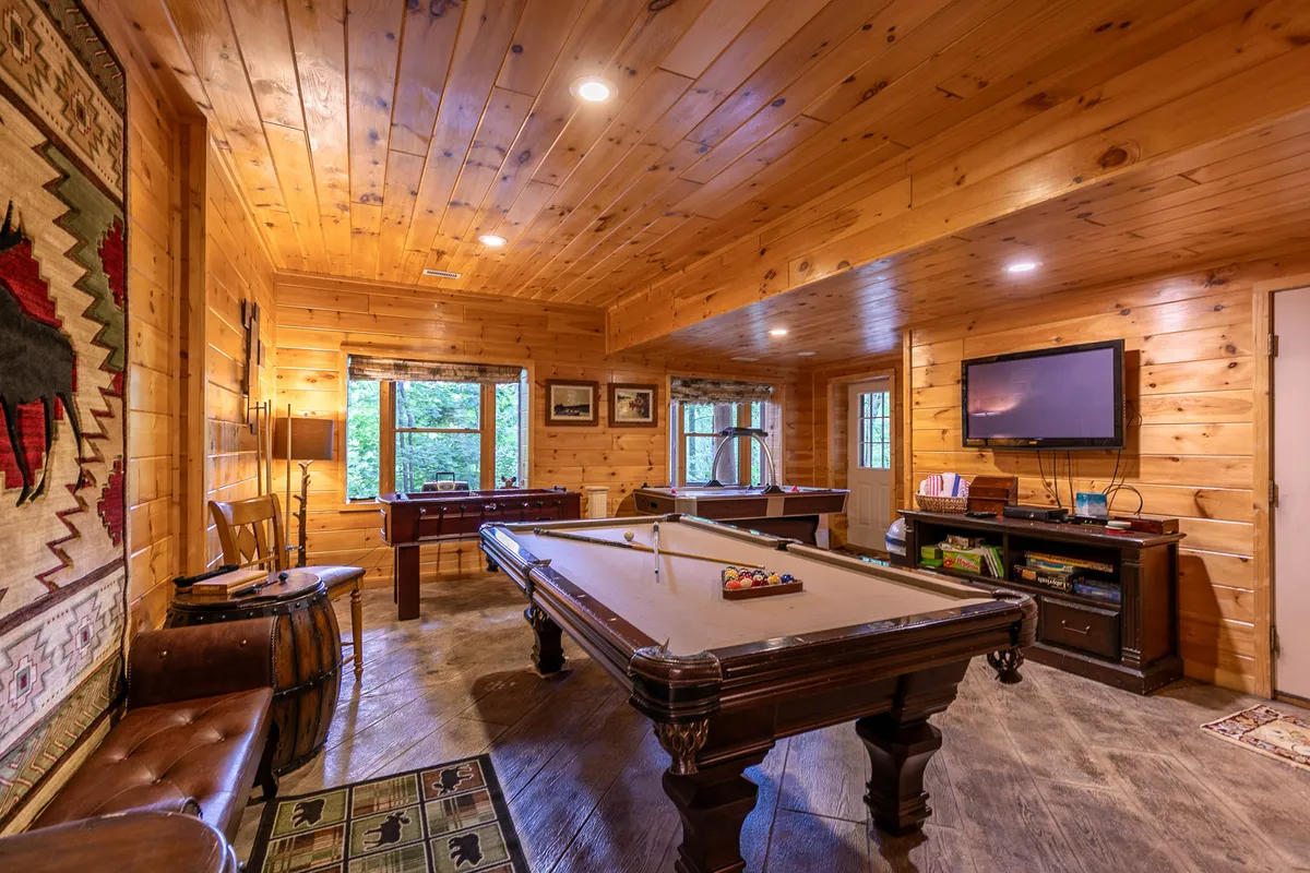 The image features a vibrant game room with a pool table as the centerpiece, surrounded by wooden walls and large windows that let in natural light. The space includes a cozy rug and a TV, creating a relaxed environment ideal for family game nights, casual competitions, and unwinding after outdoor adventures.