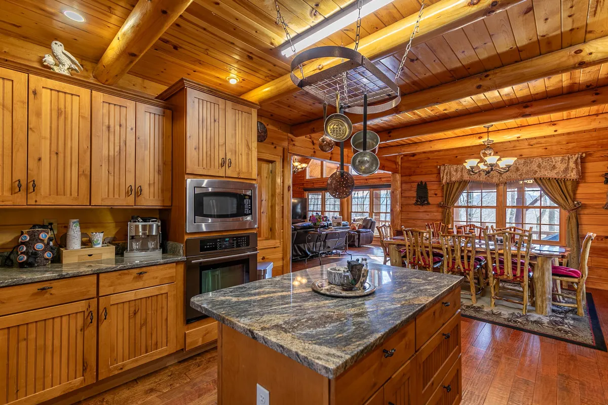 A wide-angle perspective shows the kitchen island seamlessly flowing into the adjacent dining area, surrounded by wooden cabinetry and large windows that let in natural light. The dining table is set for a group, emphasizing togetherness and easy movement between the kitchen and dining spaces.