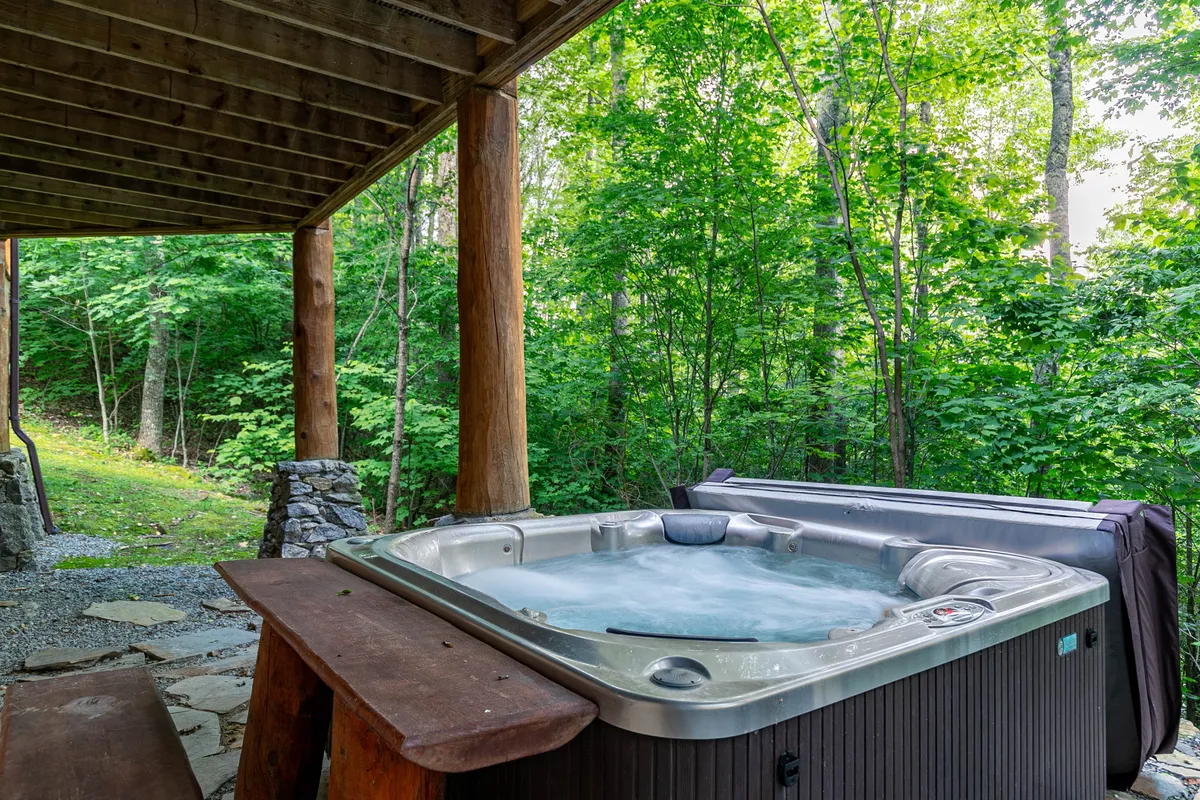 A serene outdoor scene featuring a hot tub and wooden bench surrounded by natural greenery. The hot tub area is ideal for relaxation and socializing while immersed in the peaceful outdoors, offering another dimension to the lodge’s group-friendly amenities.