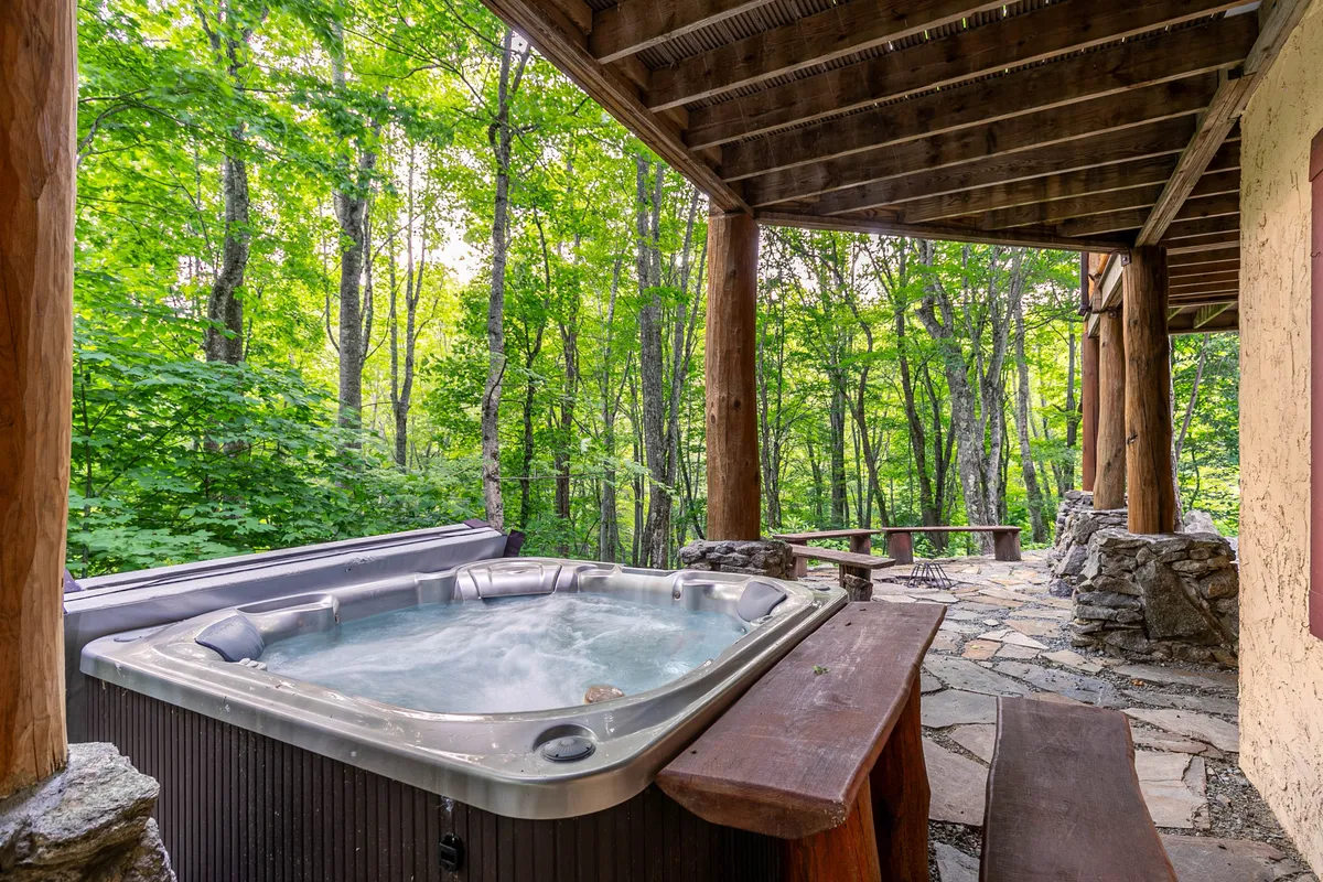 This exterior image showcases a wooden hot tub nestled among lush greenery, adjacent to a rustic log structure, with the warm glow of sunset filtering through the trees. The inviting scene suggests relaxation, social connection, and a strong connection to nature.