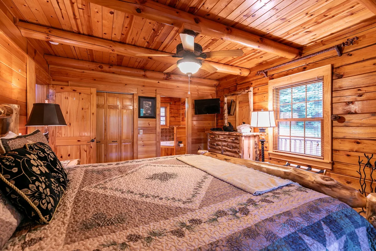 A sunlit bedroom with a wooden ceiling, a bed topped with a quilted bedspread, and large windows that frame lush outdoor views. Lamps on nightstands and a TV add modern comforts to the rustic setting, creating an inviting space for restful slumber.