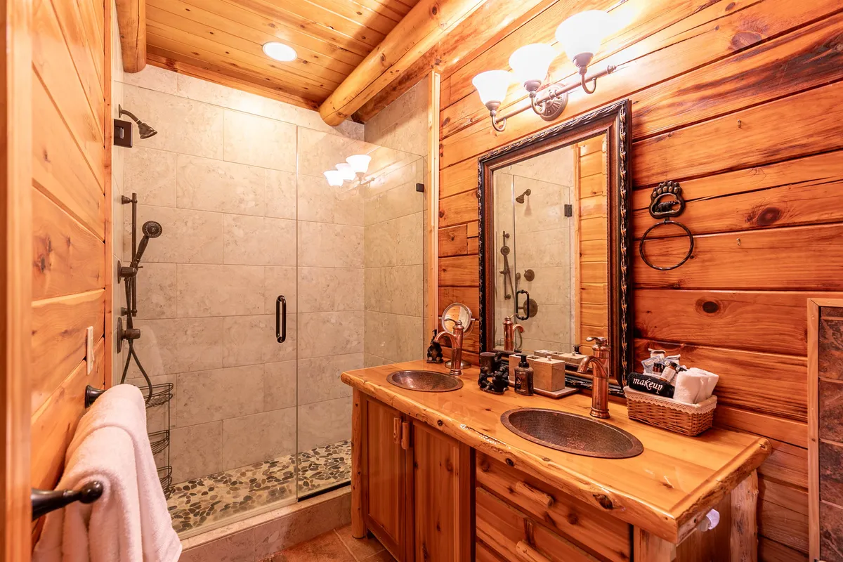 A warm, inviting bathroom with a double sink vanity, glass shower enclosure, and rustic wooden walls. The luxurious fixtures and soft lighting evoke a spa-like ambiance, ideal for relaxing self-care routines or unwinding after a day of exploration.