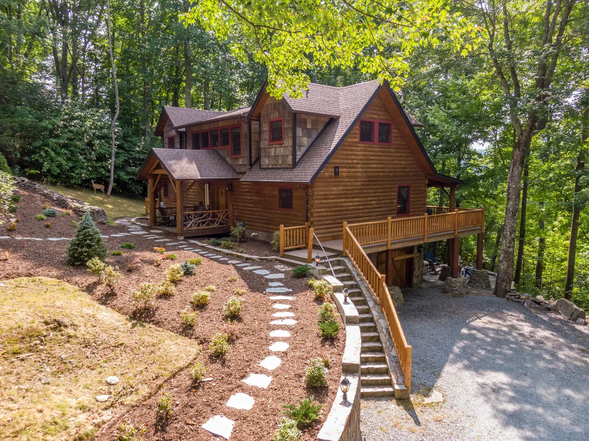 An exterior shot of a log cabin featuring a welcoming wooden deck, curved stone pathway, and plenty of space for gatherings. The setting is ideal for outdoor barbecues, stargazing, and making s'mores around a fire pit, all surrounded by greenery.