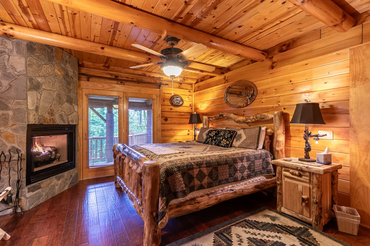 This image features a warmly lit bedroom with a rustic log bed, a stone fireplace, and handcrafted wooden furniture. Textiles with rich patterns add a cozy touch, and the room exudes an inviting, serene ambiance—ideal for relaxation and romance.
